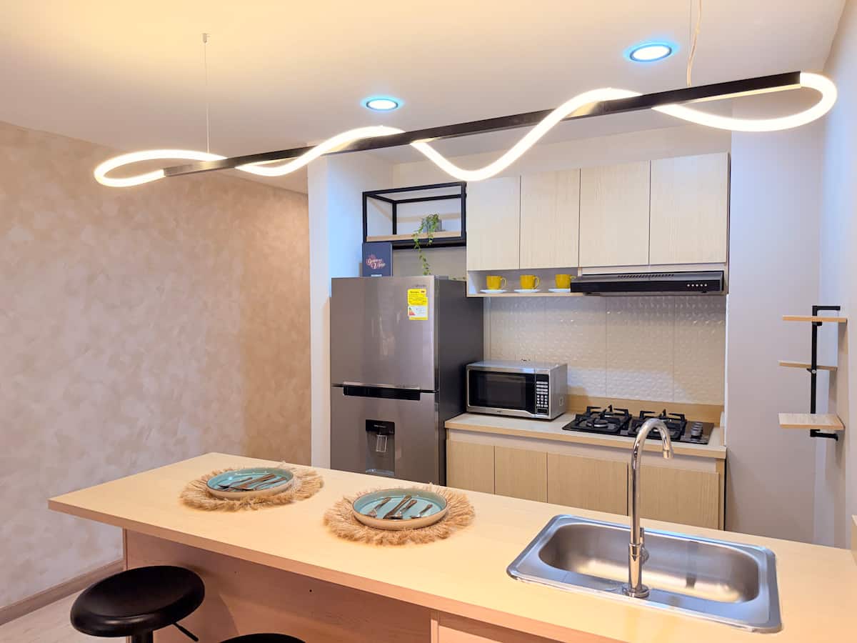 Cocina con barra de madera clara, nevera, microondas y lámpara colgante ondulada