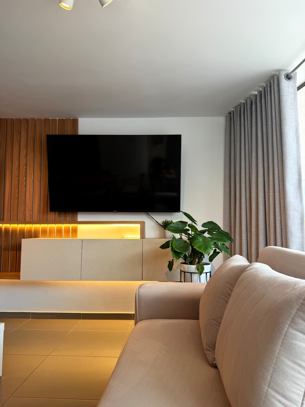 Sala con sofá beige, mueble de TV con luz cálida y cortinas grises hasta el piso