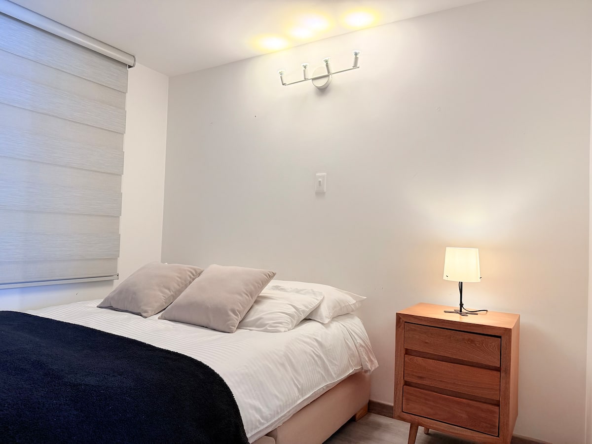 Dormitorio claro con cama doble, cobija azul oscuro y cómoda de madera con lámpara