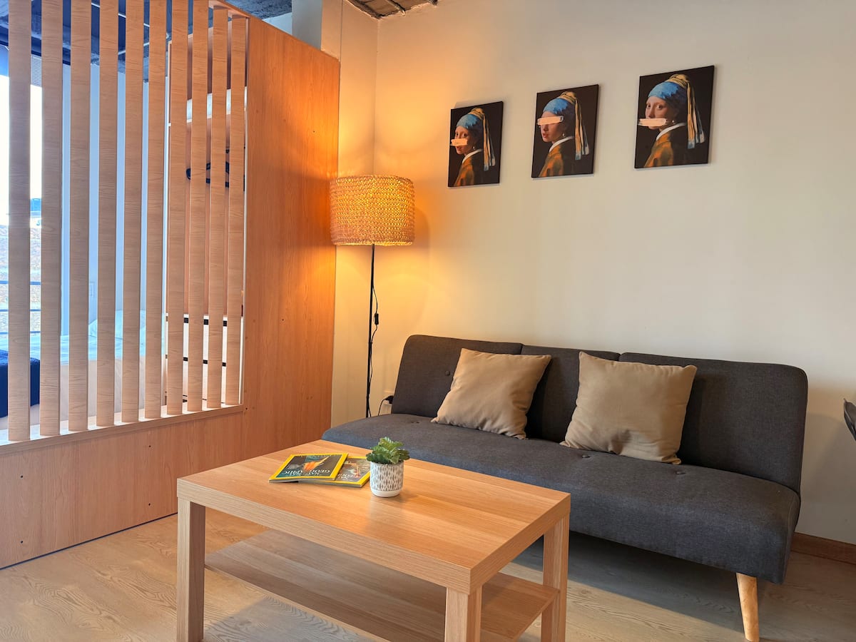 Sofá gris con cojines beige, mesa de centro de madera y tres cuadros en la pared