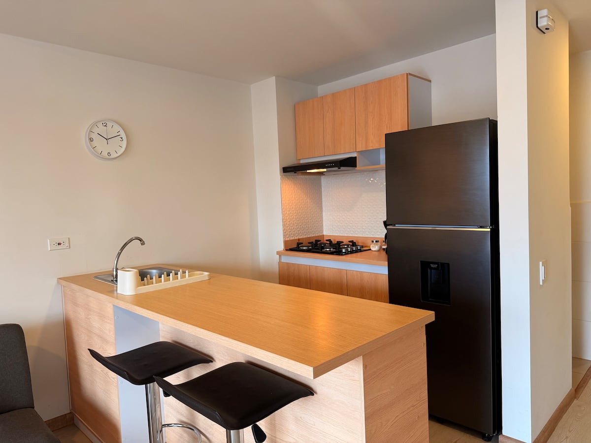 Cocina con barra de madera, dos taburetes negros, estufa a gas y nevera oscura