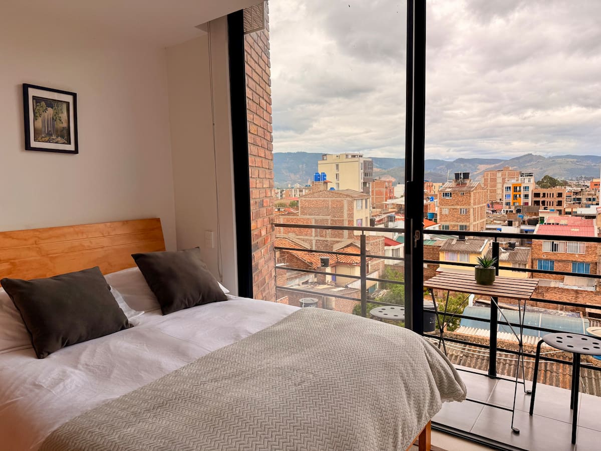 Cama doble junto al ventanal con balcón y vista a los tejados y montañas de la ciudad