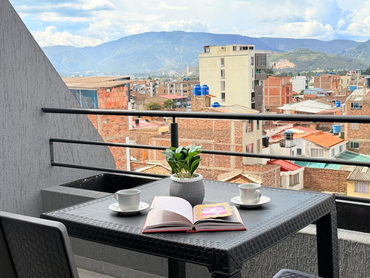 Mesa del balcón con dos tazas y un libro abierto frente a las montañas y los tejados