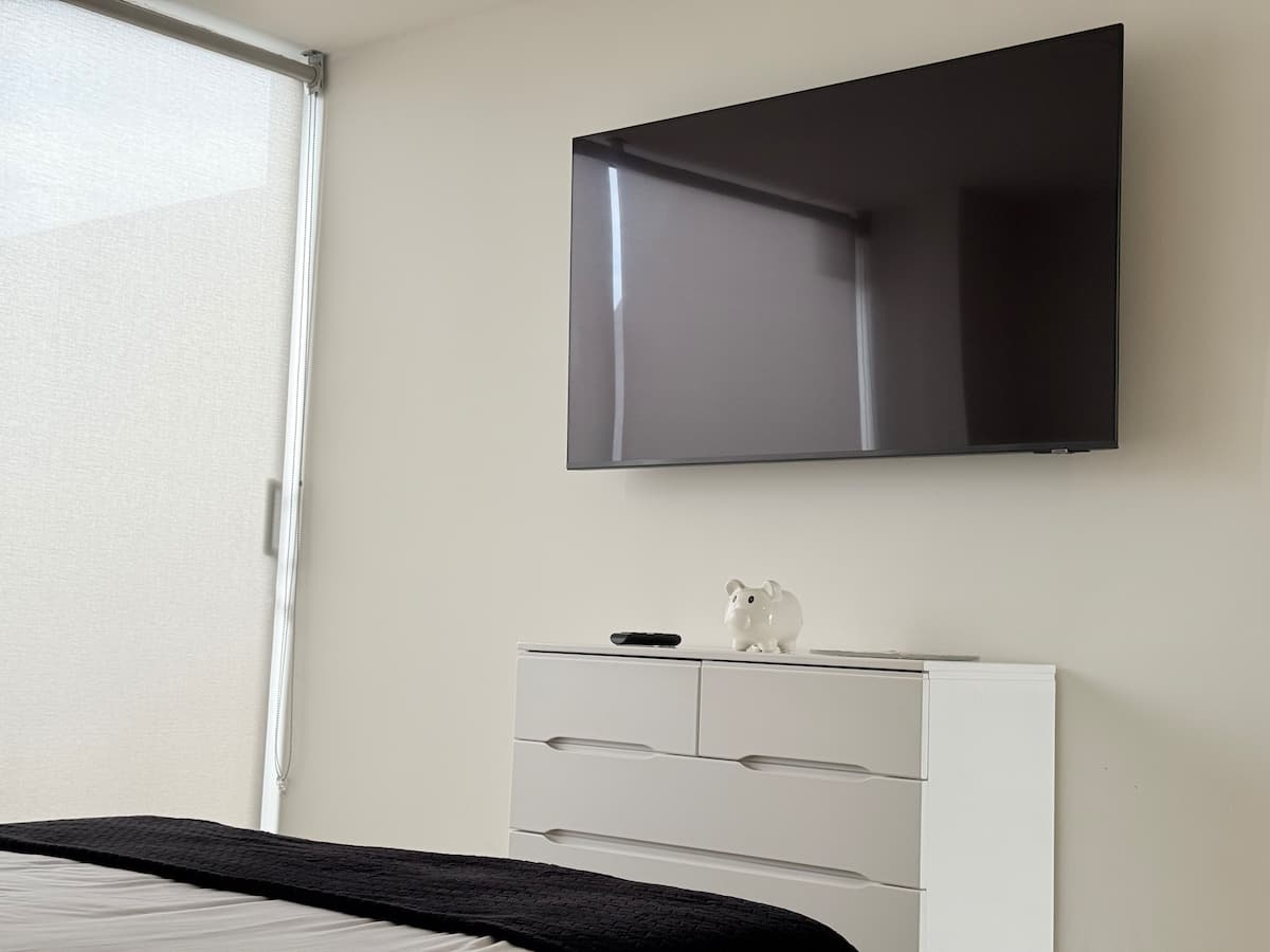 Televisor sobre cómoda blanca en el dormitorio, junto a la persiana enrollable
