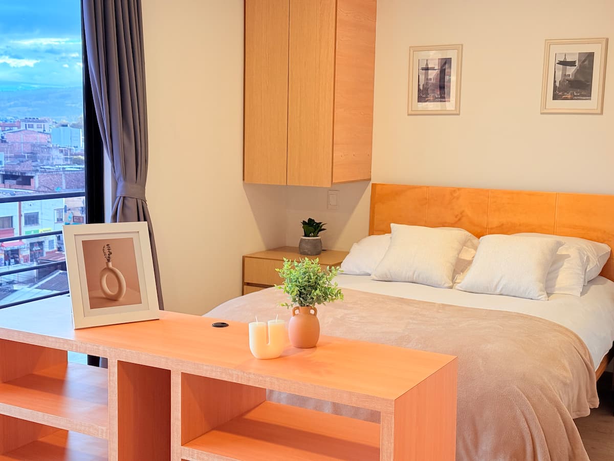 Cama doble con cabecero de madera y cuadros, junto al ventanal con vista a la ciudad